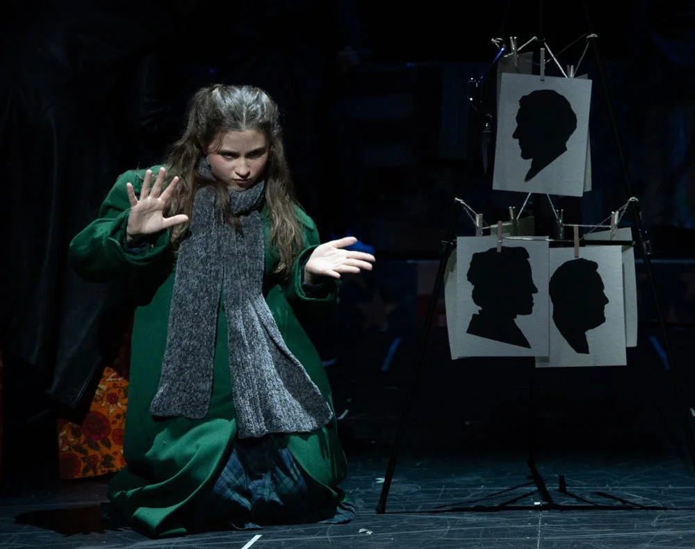 A girl dressed in green and gray is performing on stage with her hand raised, and dark silhouettes of people's profiles are displayed on panels behind her.