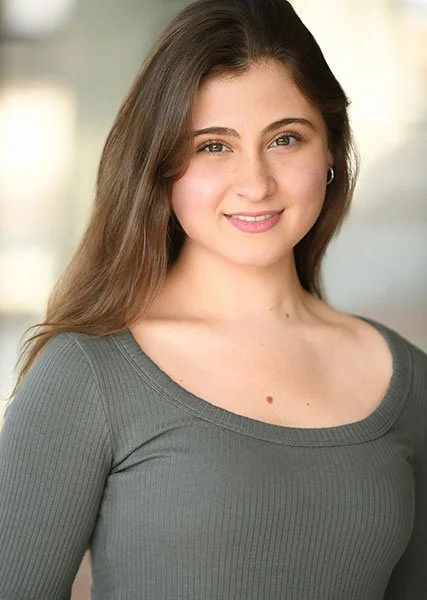 Portrait of a young woman with long brown hair, smiling, wearing a gray long-sleeve top, indoors with blurred background.