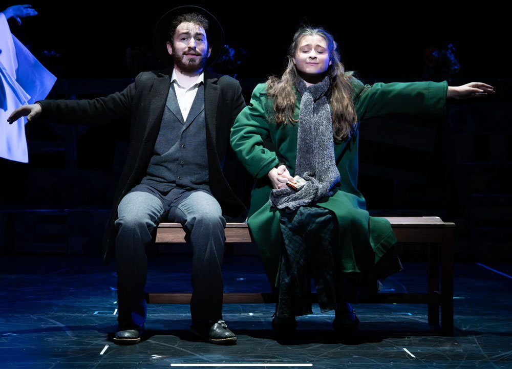 Two actors sitting on a bench on a stage, performing in a play. The actor on the left is a man with dark hair, a beard, dressed in a suit with a gray vest and tie. The actor on the right is a woman with long hair, wearing a green coat and a gray scar