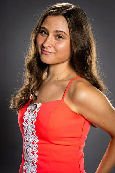 A young woman with long brown hair wearing a red sleeveless dress with white embroidery, posing against a dark gray background.