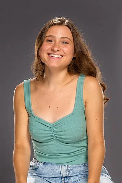 Young woman with long wavy hair smiling, wearing a turquoise sleeveless top and jeans, sitting against a gray background.