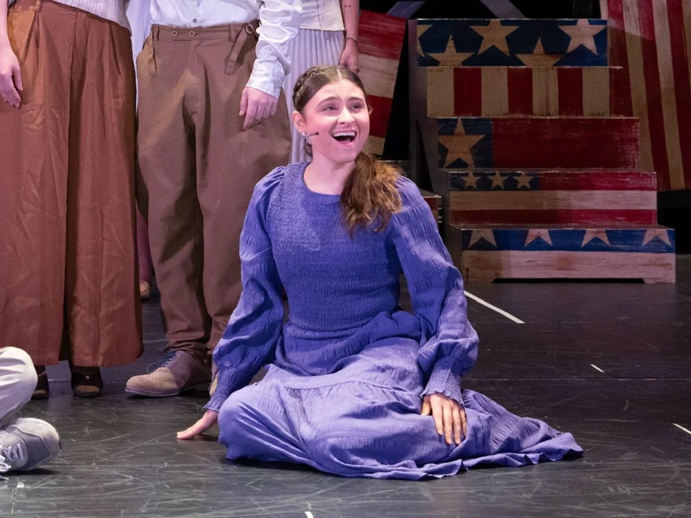 Young woman with long hair sitting on the stage floor, smiling and singing, wearing a purple dress, surrounded by other people in period costumes and patriotic decorated stairs in the background.