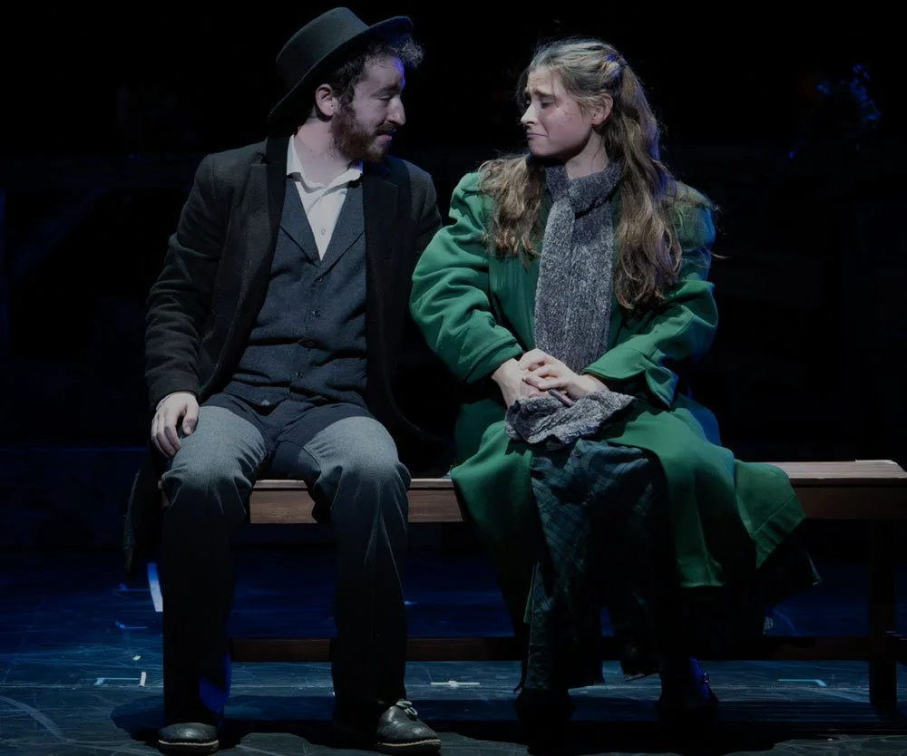 A man and woman sitting on a bench on stage, engaged in a serious conversation, with dark lighting and theatrical setting.