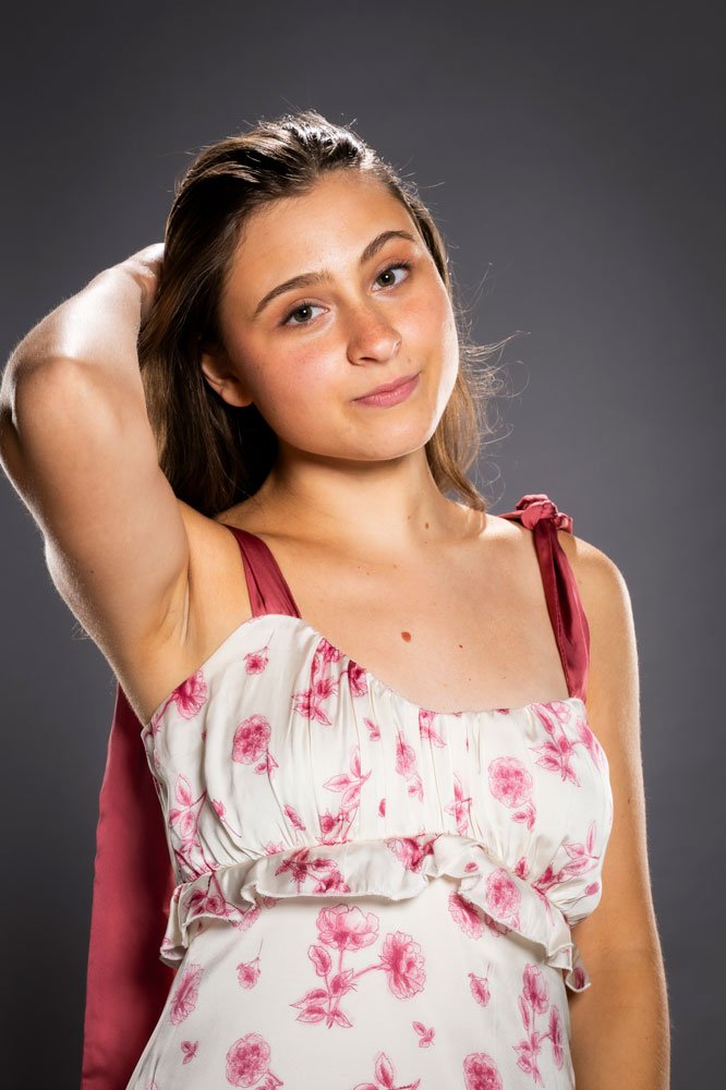 A young woman with long brown hair, wearing a white dress with pink floral pattern and maroon straps, stands against a dark gray background, looking at the camera with a slight smile and one arm raised behind her head.