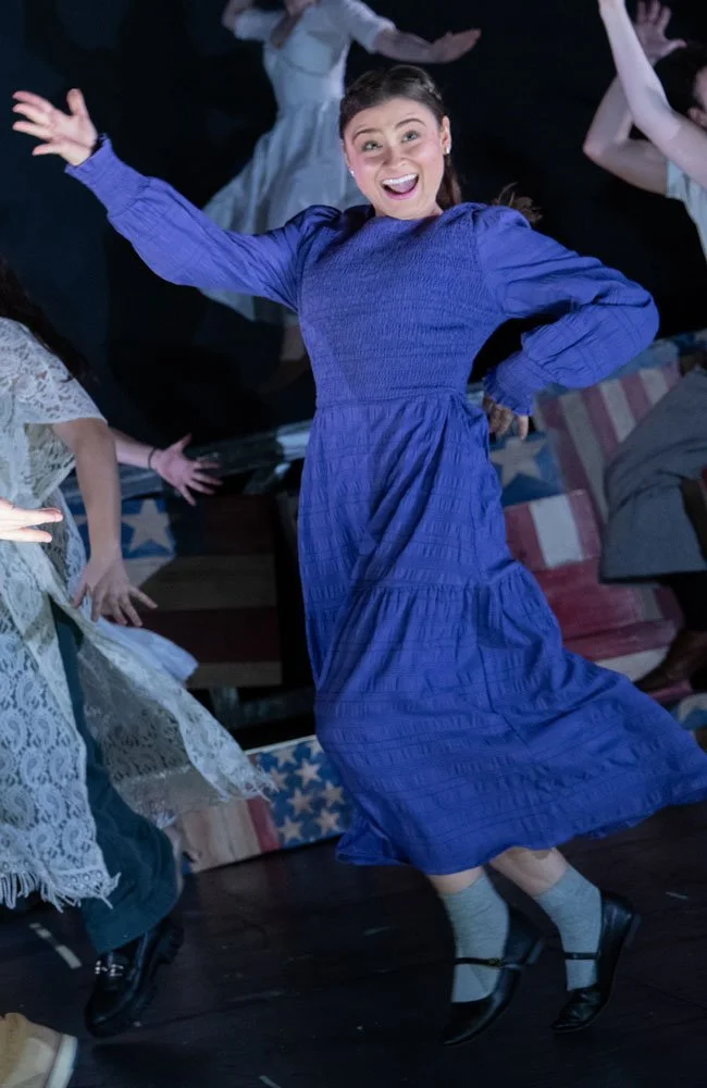 A woman in a blue dress dancing and smiling, surrounded by other dancers, on stage with American flag-themed decor.