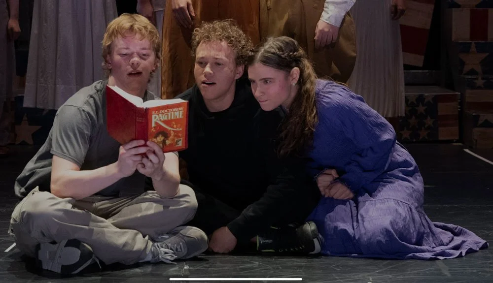 Three young actors performing on stage, sitting on the floor, with a book in hand, in a theatrical production.