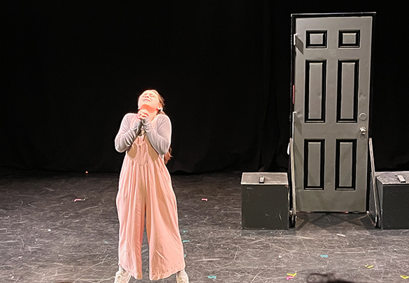A young girl in a pink jumpsuit stands on a stage with her hands clasped and looking upward. There is a closed door and two black boxes on each side in the background.