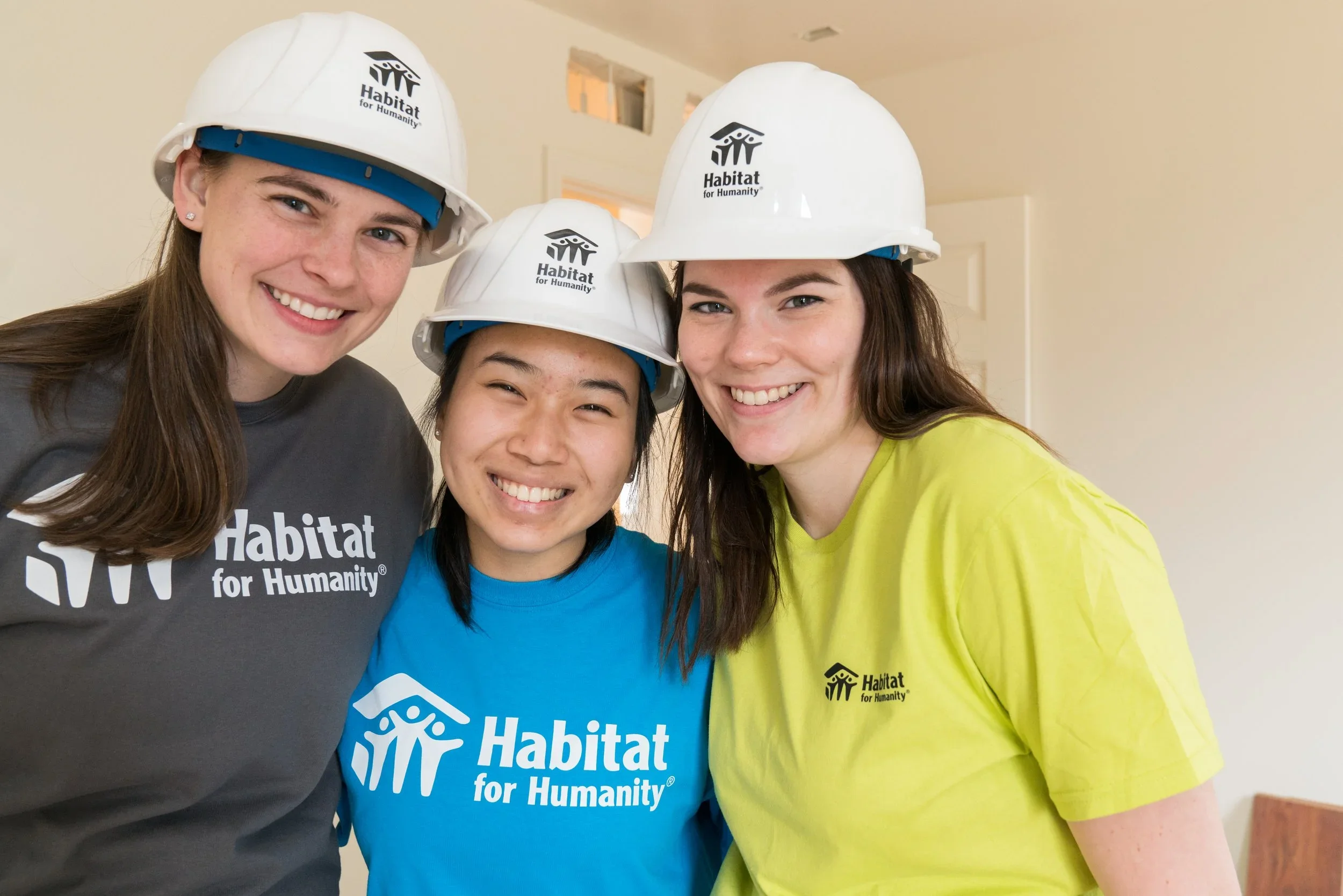 Three women construction volunteers wearing Habitat for Humanity hard hats and T-shirts, smiling indoors.
