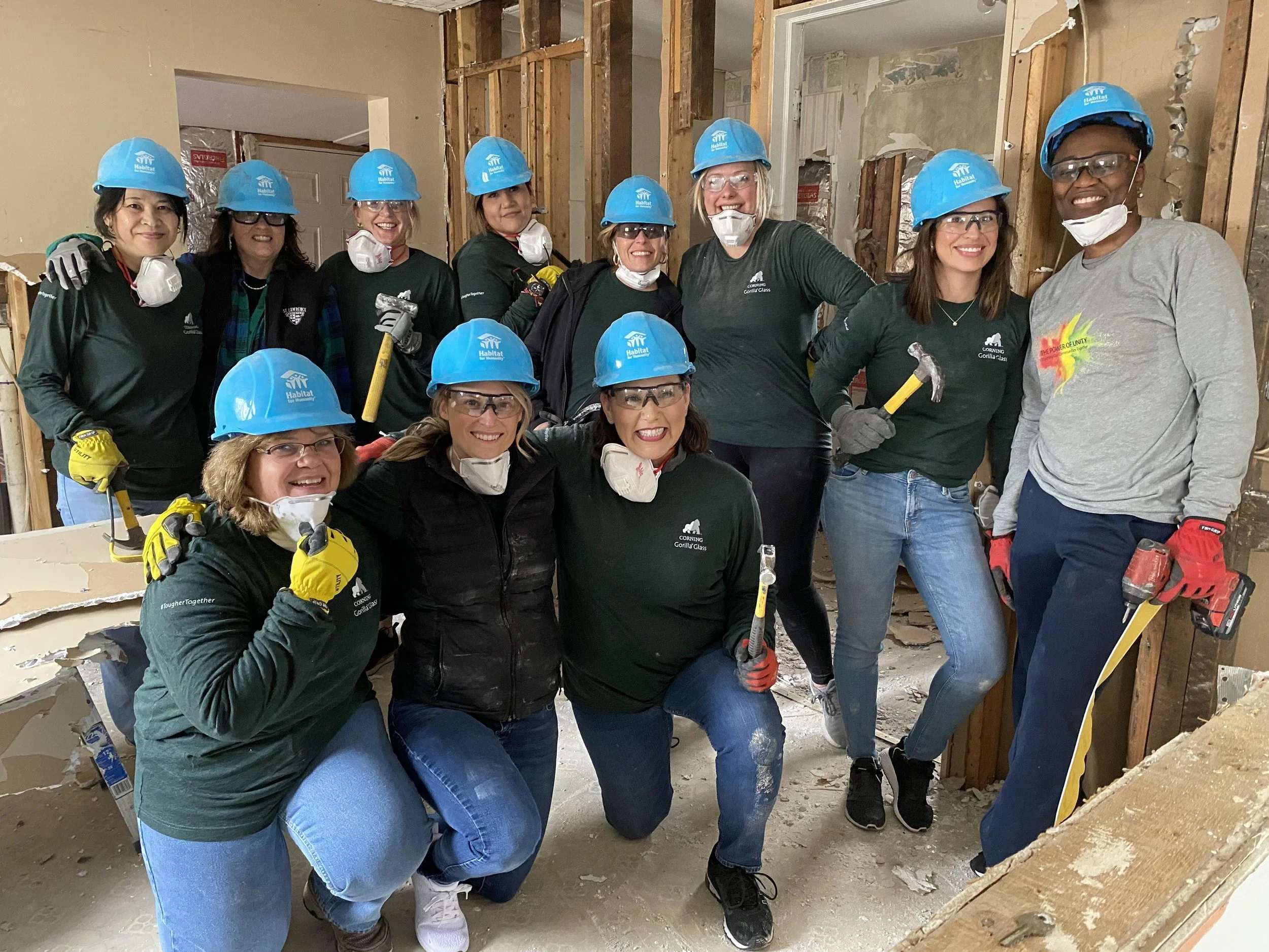 Group of volunteers in construction gear, including blue hard hats, safety glasses, and gloves, working together on a home renovation project, smiling for the camera.