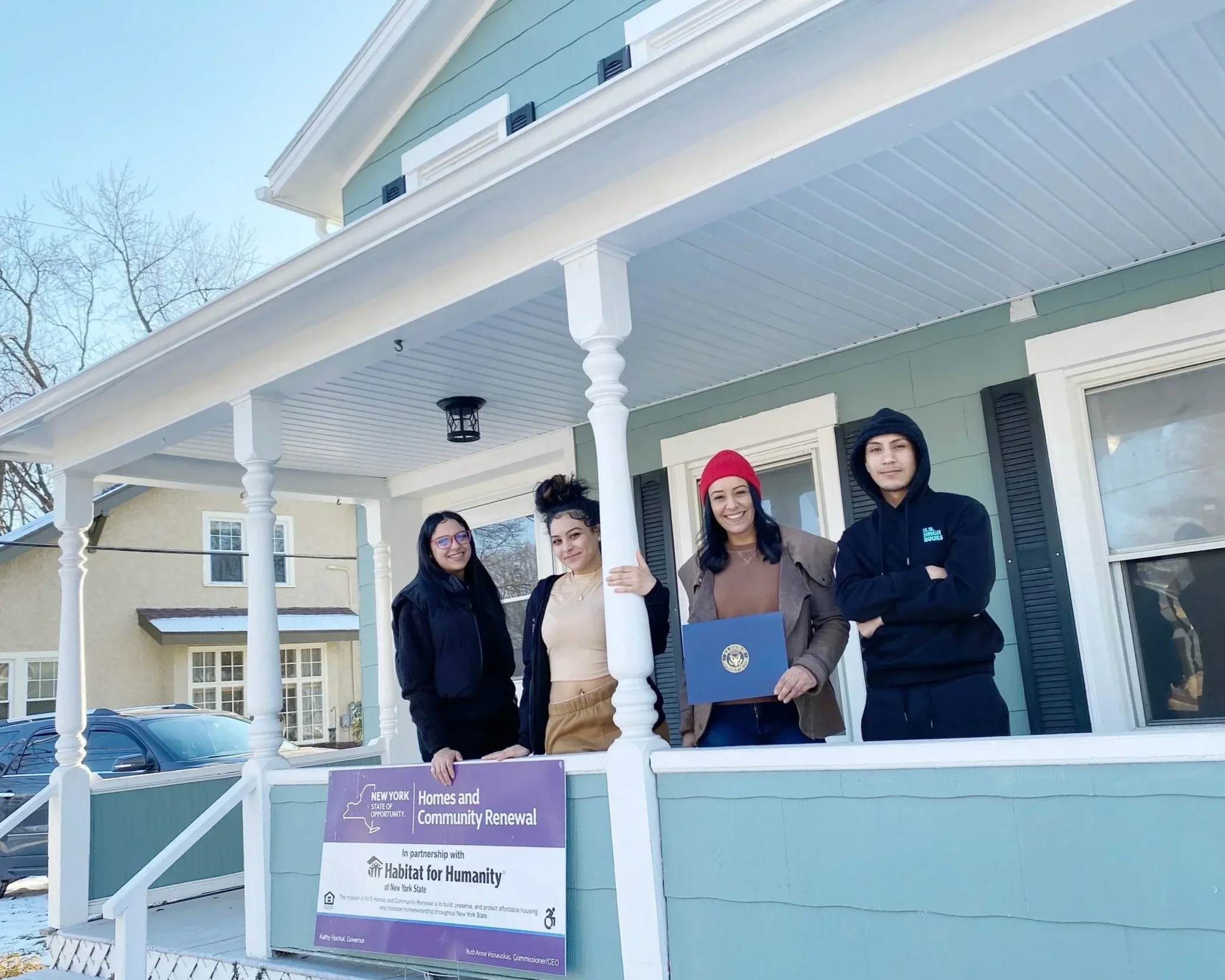A group of new Habitat for Humanity homeowners standing on a porch holding signs, participating in a community renewal event, with a banner and a residential neighborhood in the background.