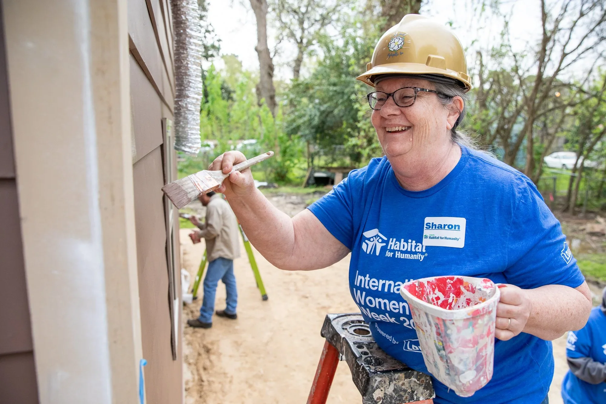 A woman construction volunteer wearing a blue shirt with Habitat for Humanity and International Women’s Week text painting a house exterior with a brush. She is smiling and holding a paint bucket, with a man and ladder visible in the background.