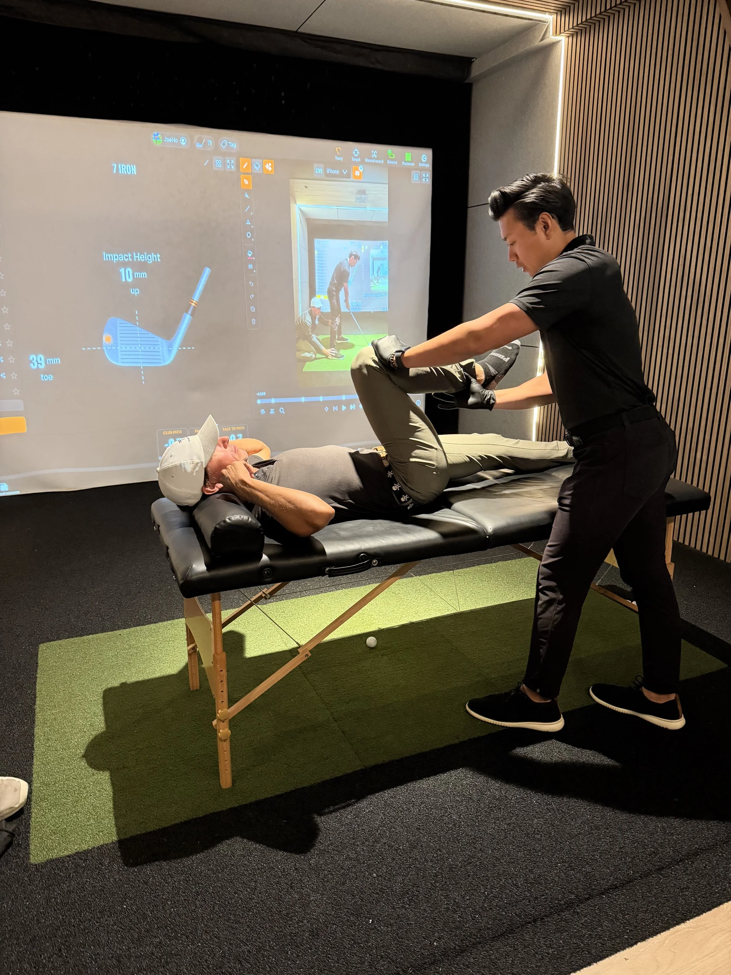 A person lying on a massage table receiving a golf swing analysis from a professional. There is a large screen in the background displaying golf club data and a video of a golf swing.
