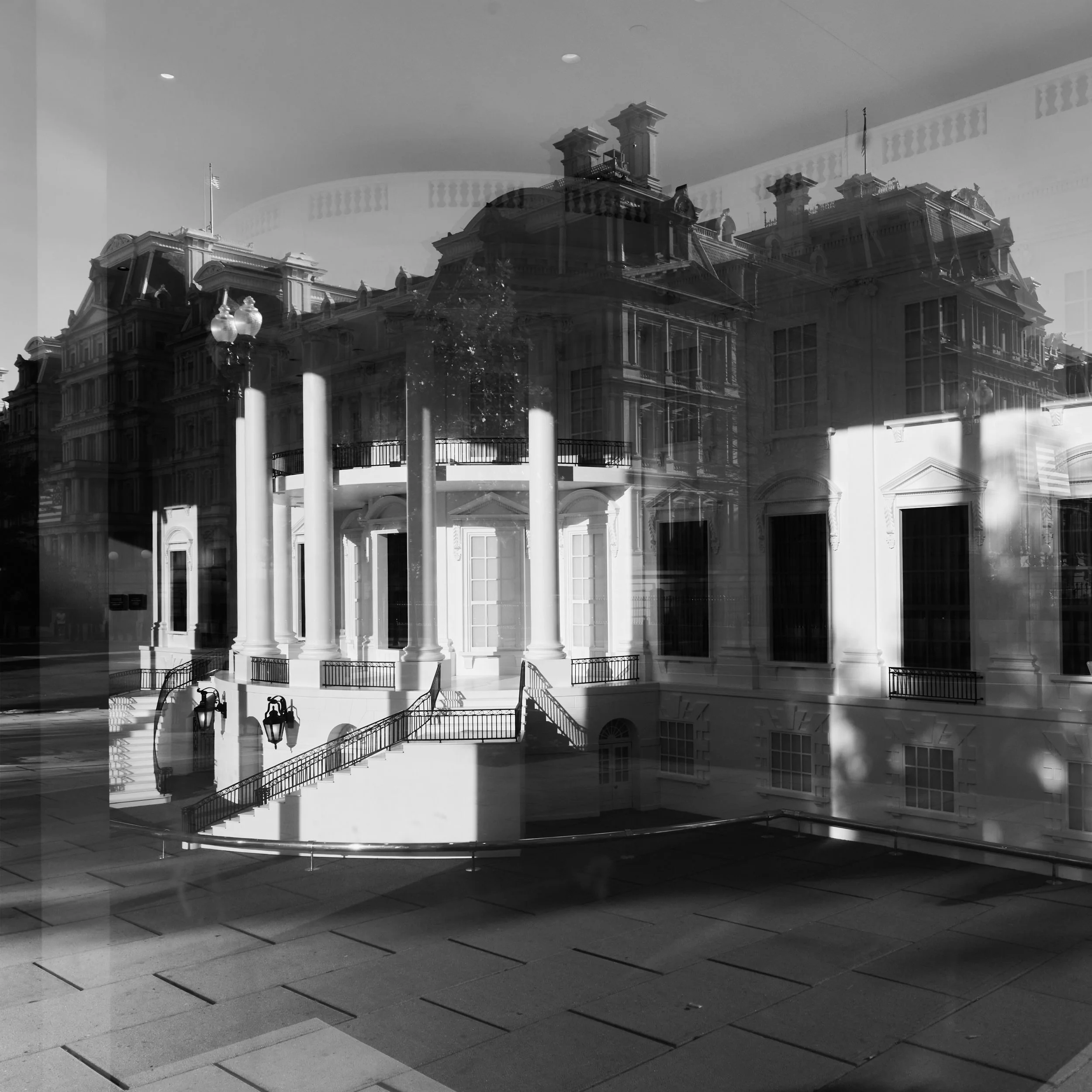 A black and white photograph of a large, ornate building with columns, decorative railings, and multiple chimneys on the roof, seen through a reflective glass window, with a staircase and street lamps in the foreground.