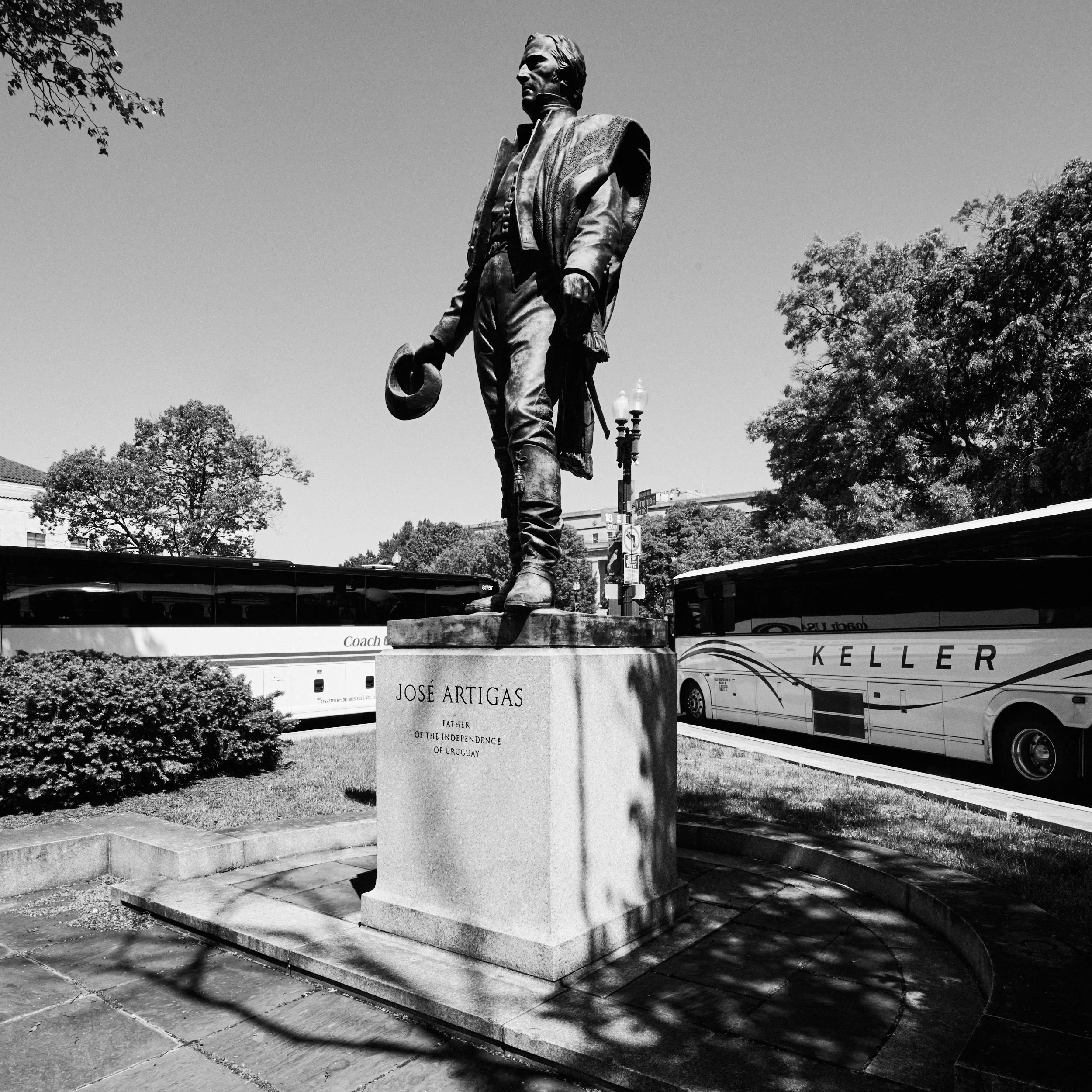 Black and white photo of a statue of a man holding a wide-brimmed hat, standing on a pedestal in an outdoor park with trees in the background.