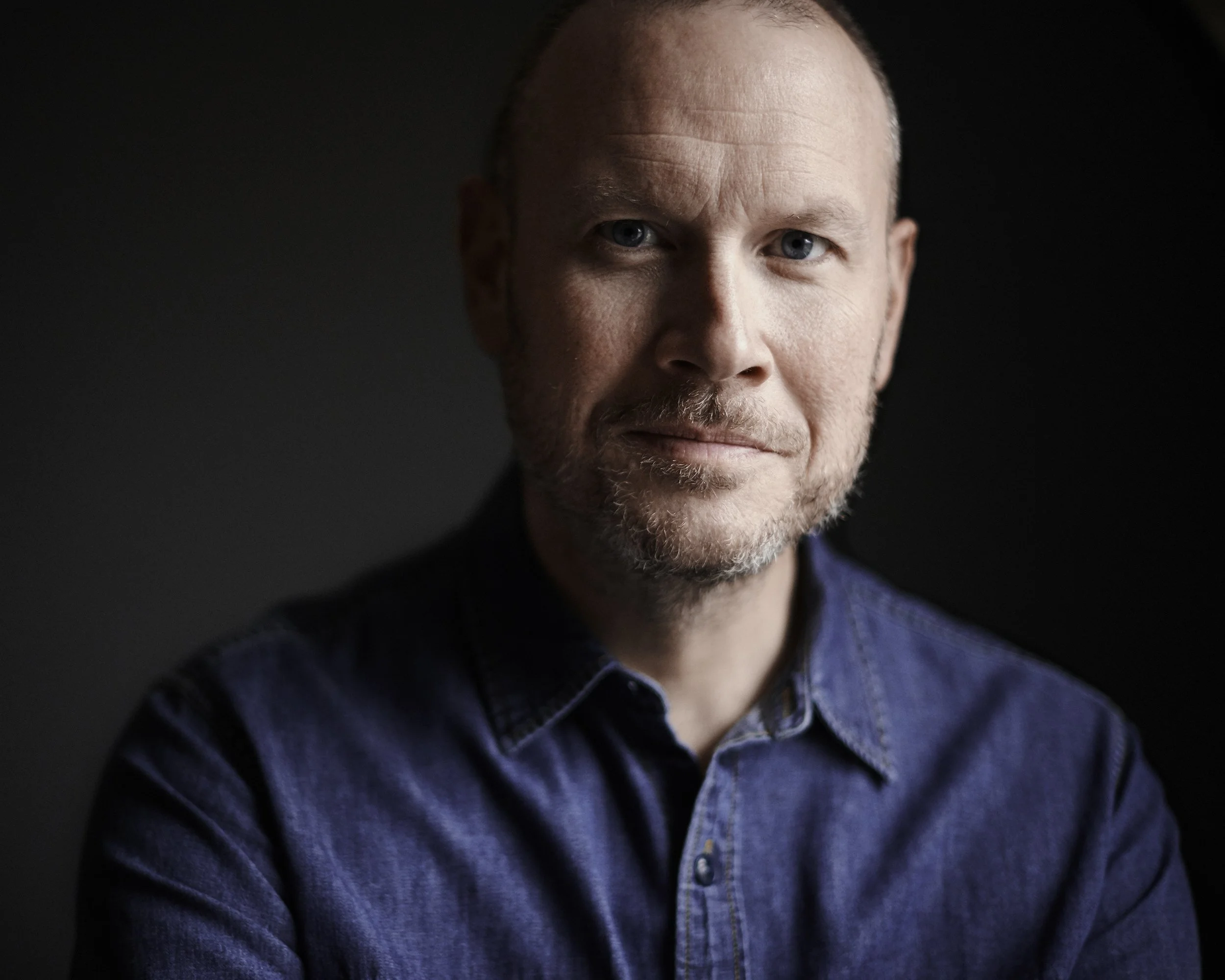 A close-up portrait of a middle-aged man with short light brown hair, a trimmed beard, and blue eyes, wearing a blue denim shirt, against a dark background.