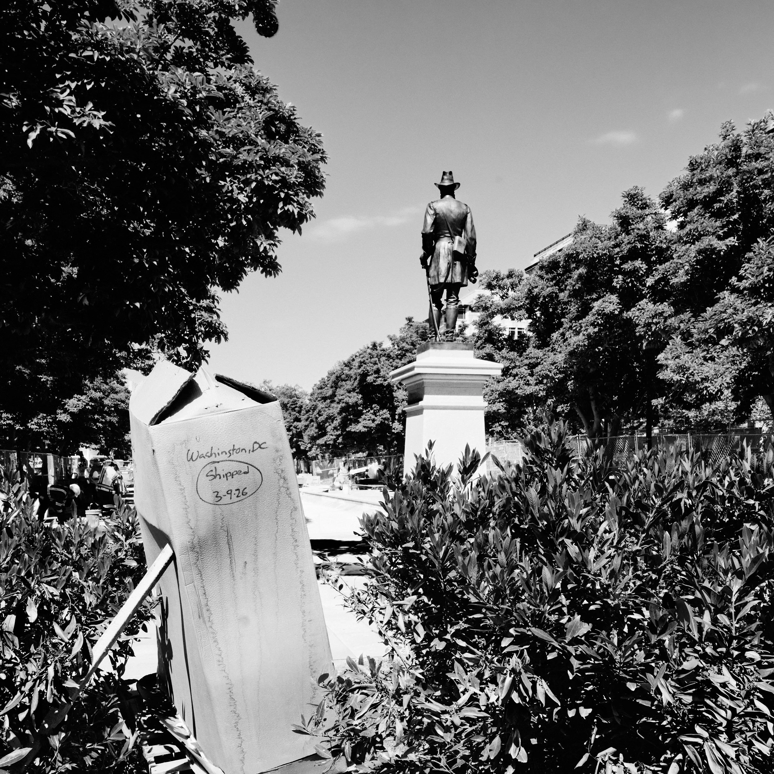 A street scene with a statue on a pedestal, trees with dense foliage surrounding it, and a sign in the foreground indicating shipment from Washington, D.C. The photo is in black and white.