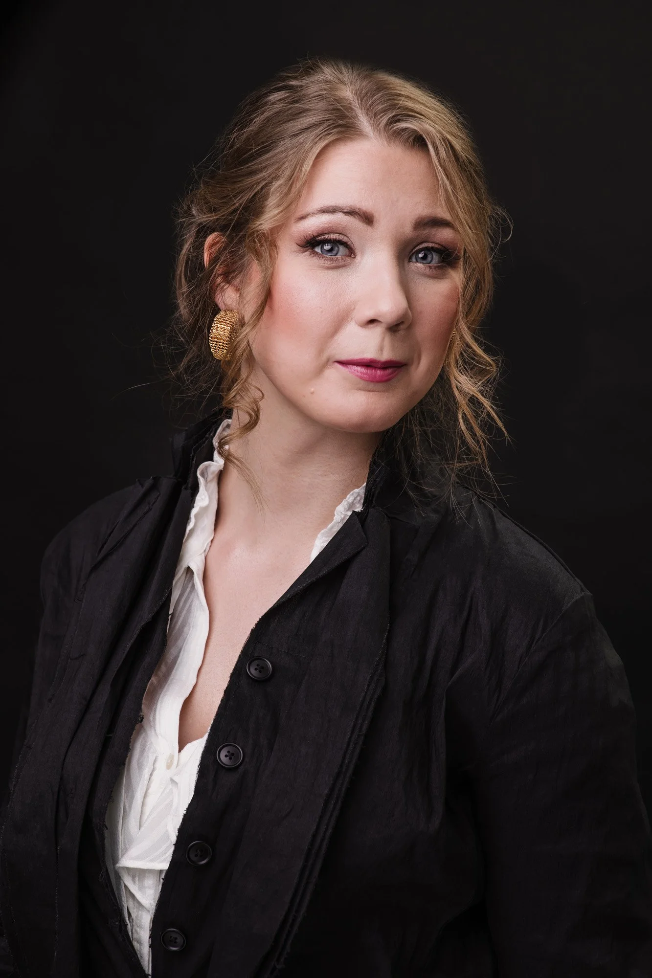 Close-up portrait of a woman with blonde hair and blue eyes, wearing a black jacket over a white blouse and gold earrings, against a black background.