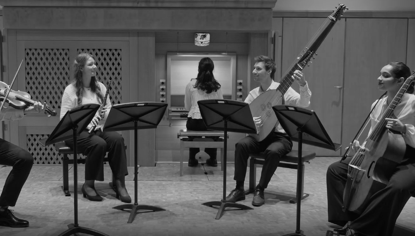 A group of musicians playing classical instruments in a recording studio, including a violin, clarinet, guitar, and cello, with a person at the piano in the background.