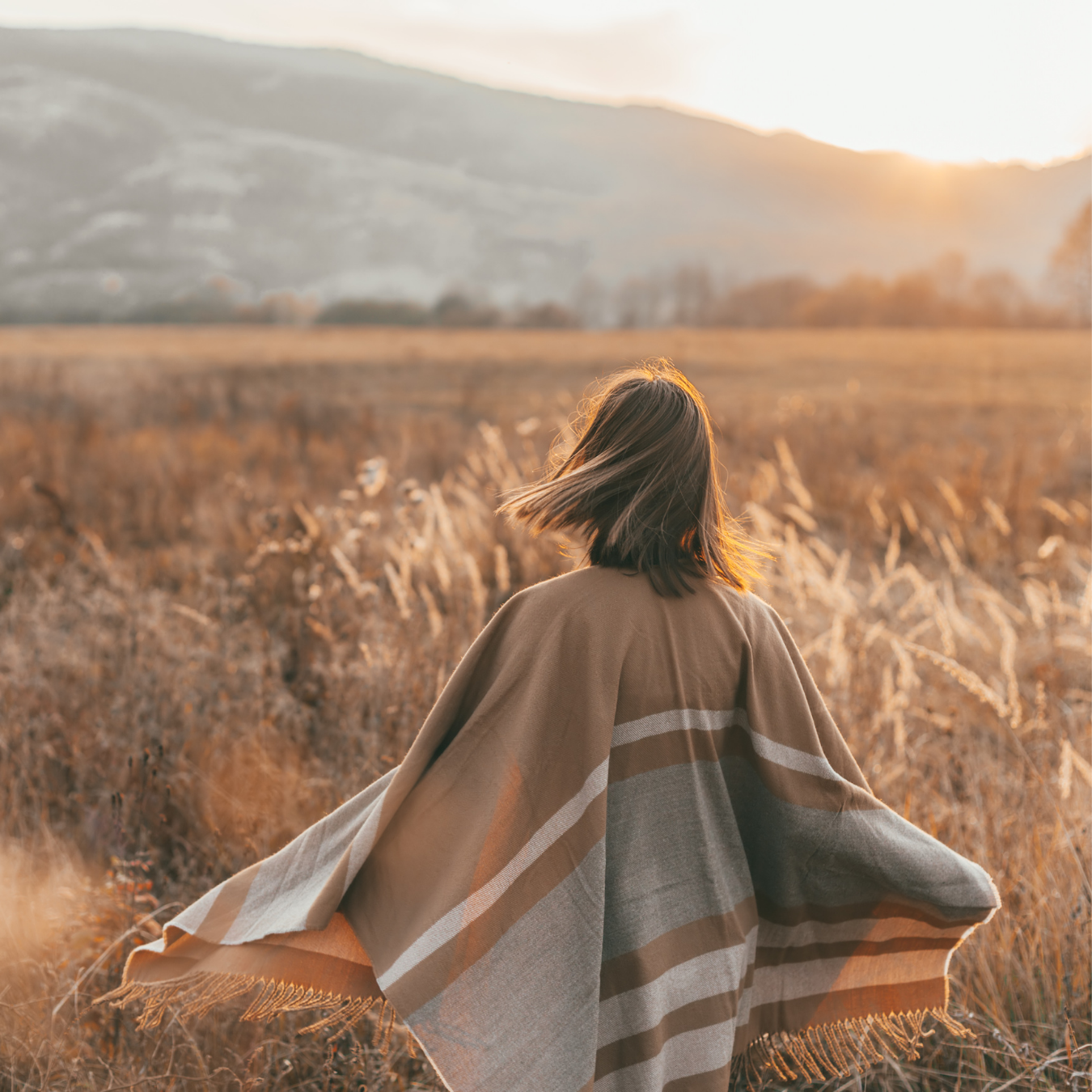 A person with shoulder-length hair walking through a field of tall dry grass during sunset, wearing a broad striped shawl.