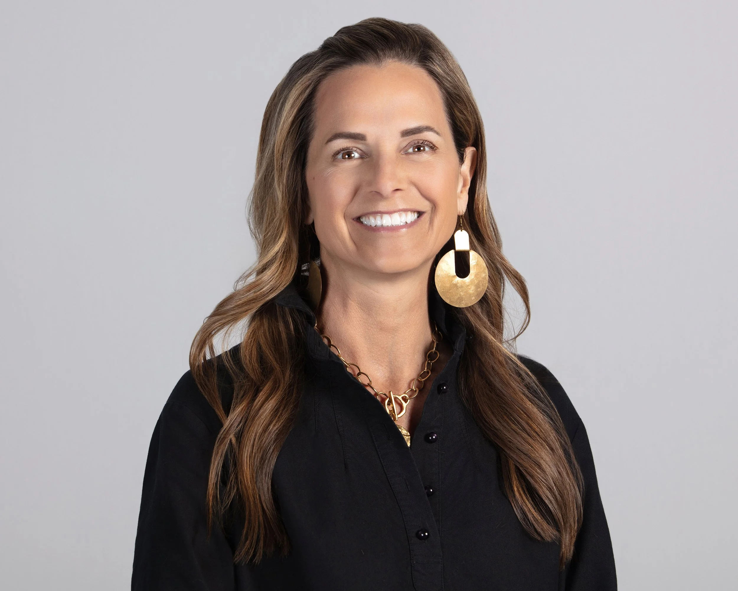 A woman with long wavy brown hair, wearing a black button-up shirt and large gold earrings, smiling against a plain light gray background.