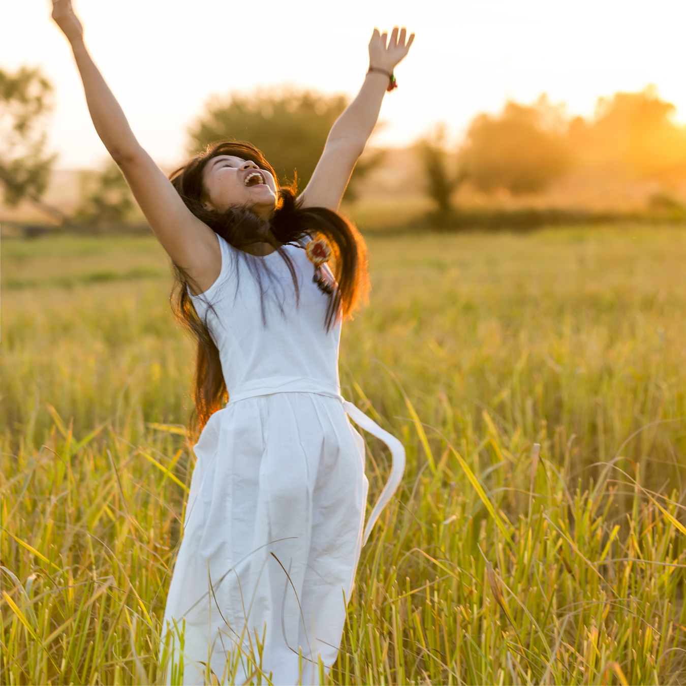 A woman in a white dress with a feather earring standing in a field of tall grass during sunset, smiling and raising her arms joyfully.