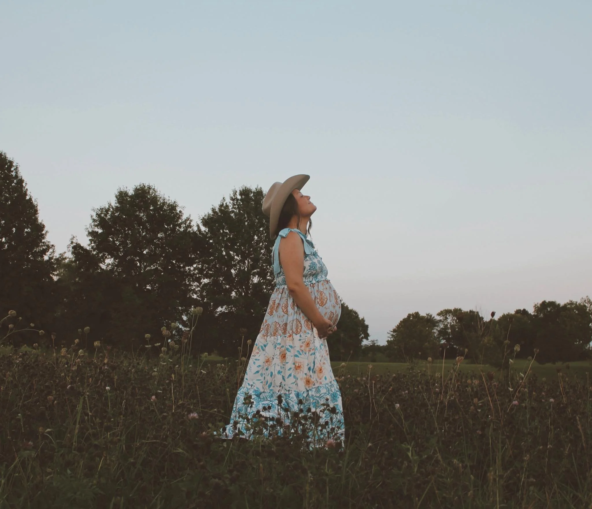 Pregnant woman in a floral dress and wide-brimmed hat standing in a field of wildflowers, looking up at the sky with trees in the background during dusk.