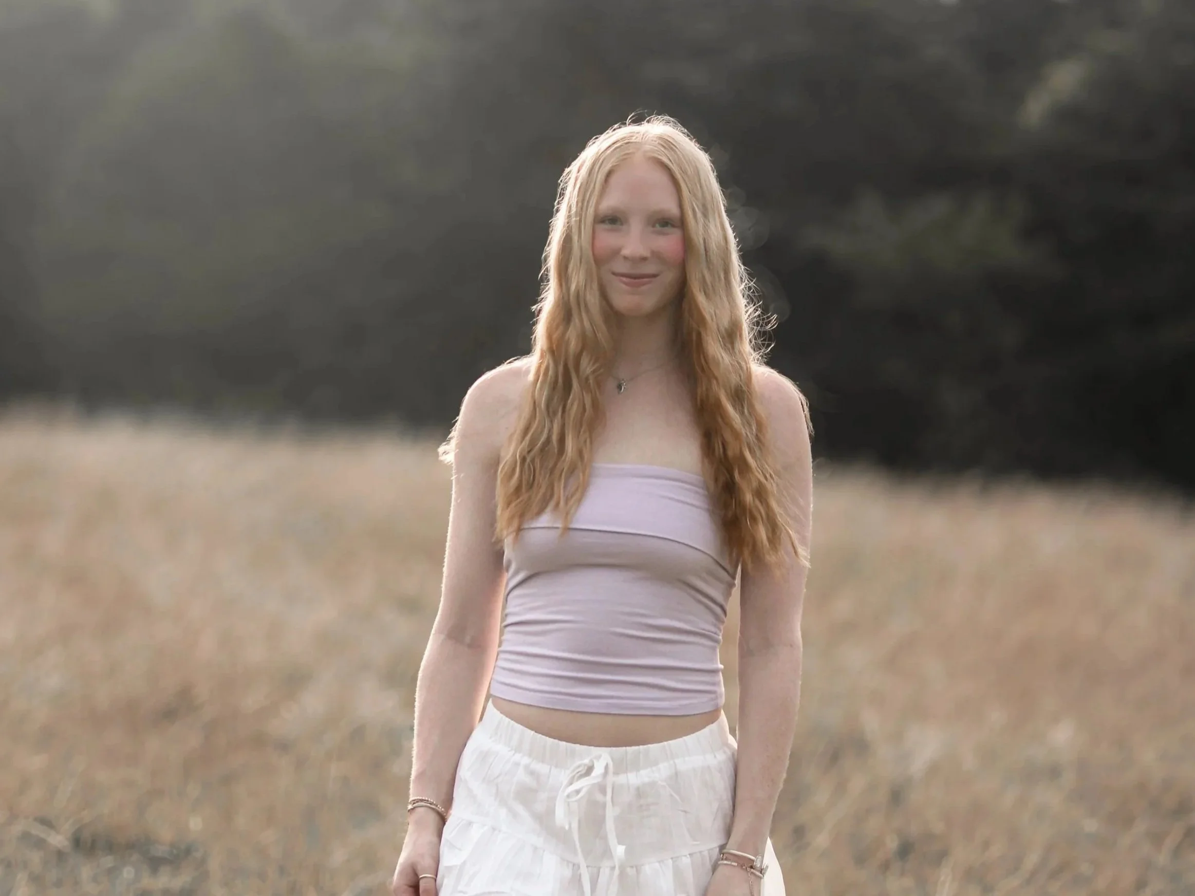 A woman sitting on a blanket outdoors in a park holding a baby in her lap, looking at the baby with affection. The scene is surrounded by trees and grass.