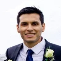 Portrait of a man with dark hair, smiling, dressed in a suit and tie, outdoors against a cloudy sky background.