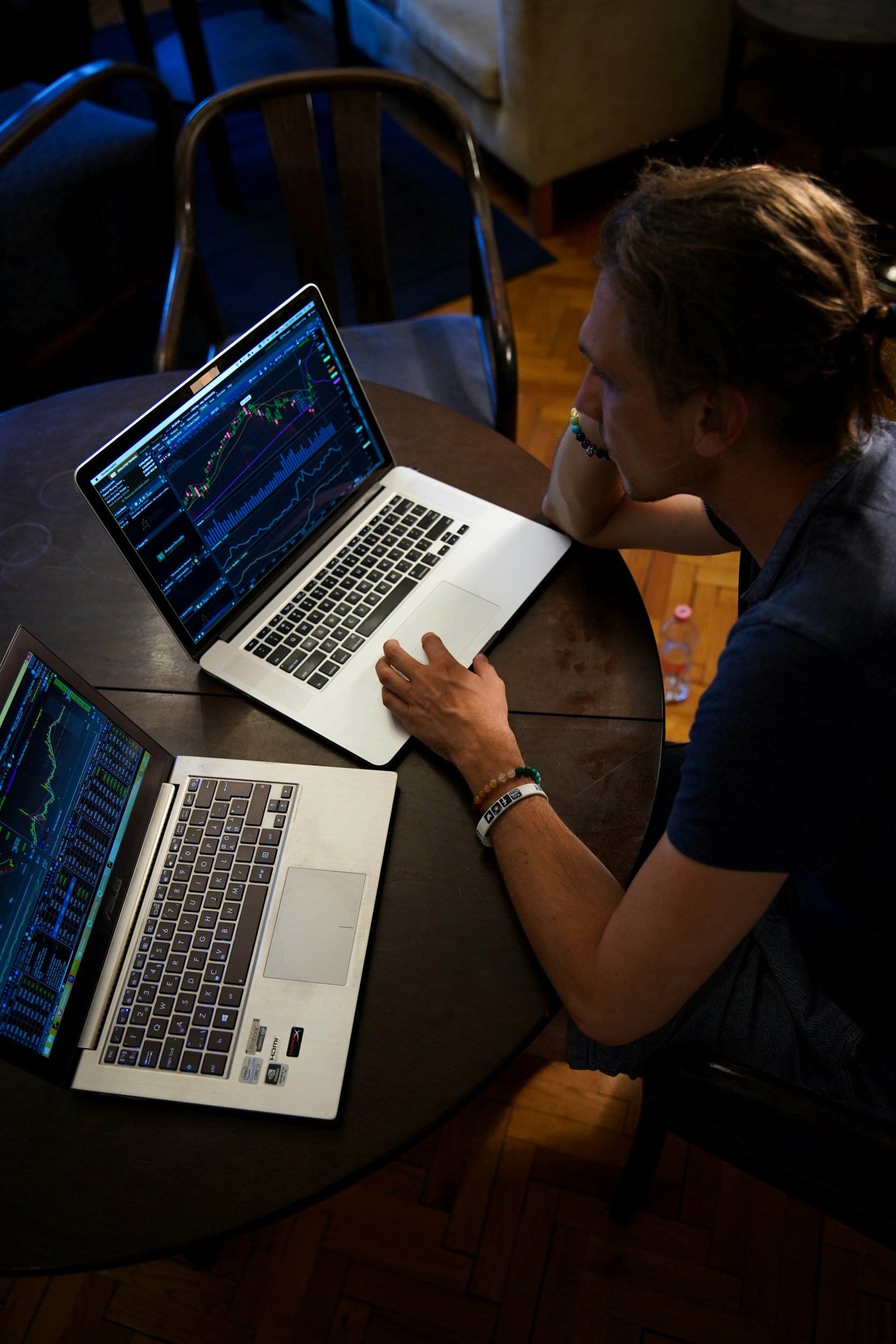 Person looking at two laptops displaying stock trading charts on a round wooden table.