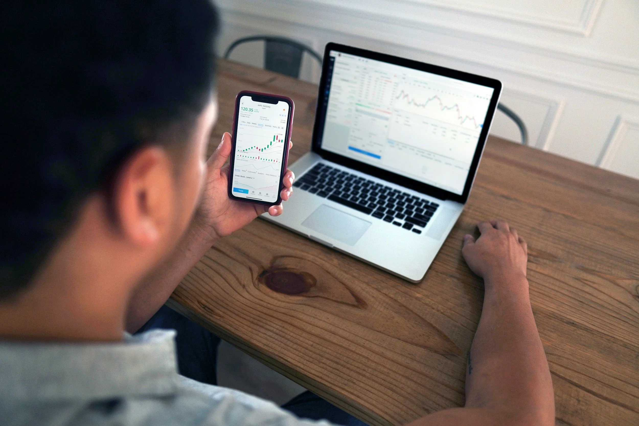 A man sitting at a wooden table holding a smartphone displaying a stock trading app, with a laptop open in front showing stock charts and trading data.