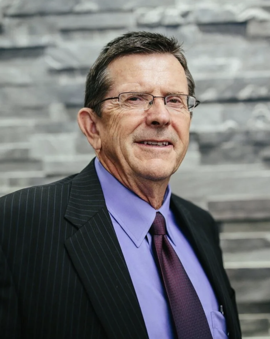 A middle-aged man wearing glasses, a dark suit with pinstripes, a purple shirt, and a maroon tie, standing in front of a gray stone wall.