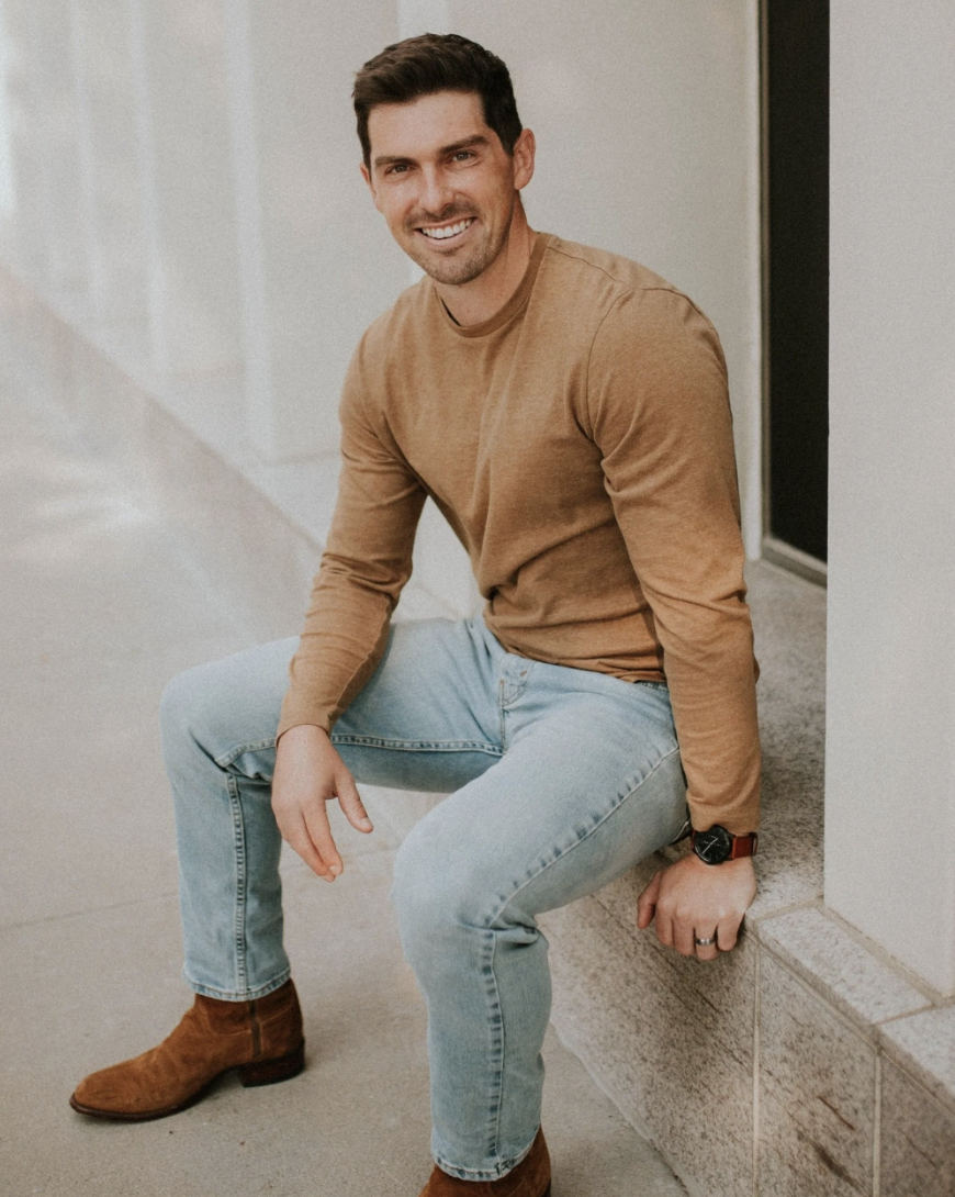 A smiling man sitting on a concrete ledge on a sidewalk outdoors. He has dark hair, is wearing a brown long-sleeve shirt, light blue jeans, and brown boots. He is looking at the camera. Zach Lind of Lind Financials.