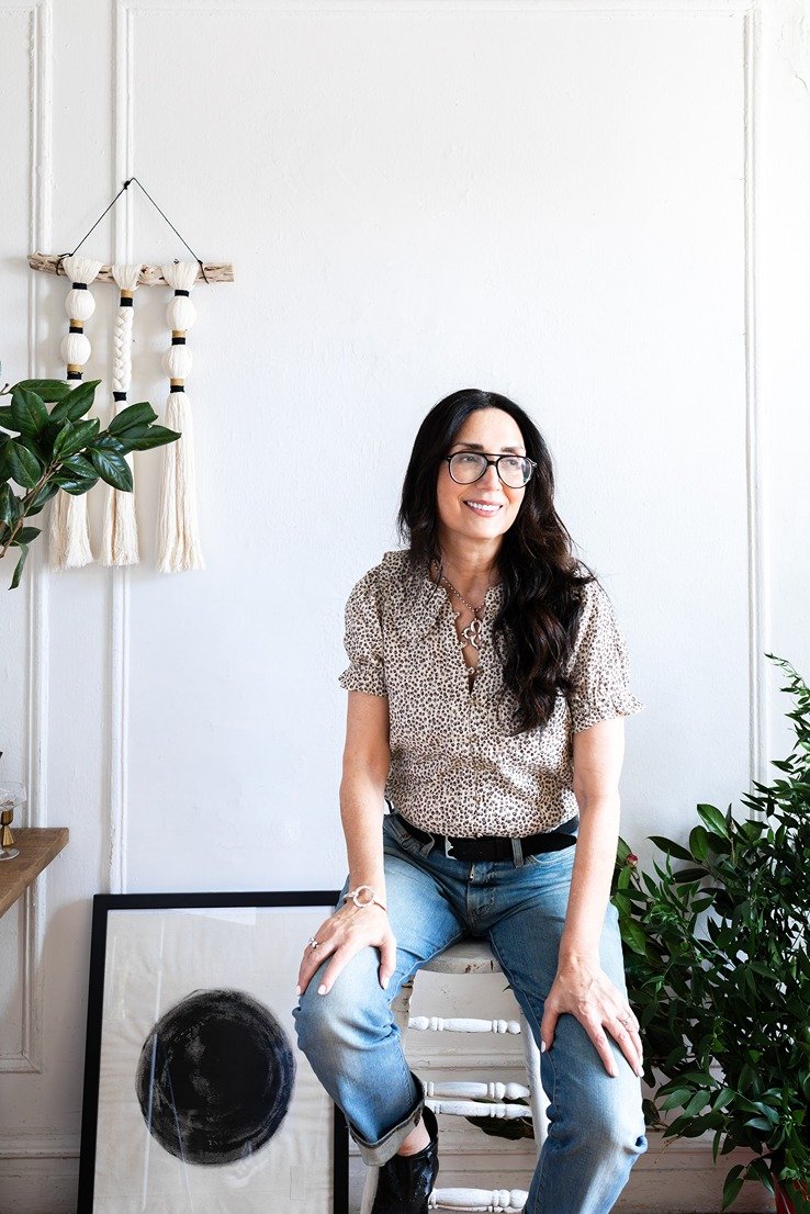 Woman with long dark hair and glasses sitting on a white chair in a room with plants and artwork, smiling.