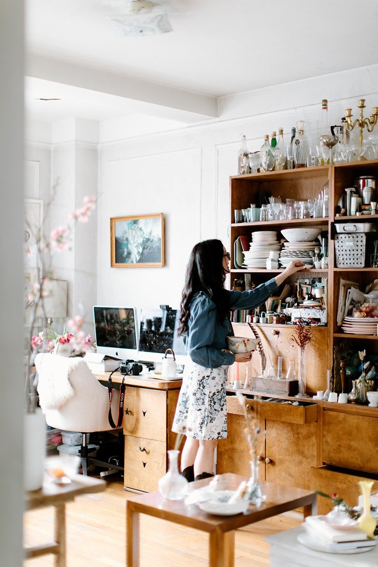 Woman looking at glassware on wooden shelf in a cozy, well-lit room with a computer desk, decorative items, and artwork.