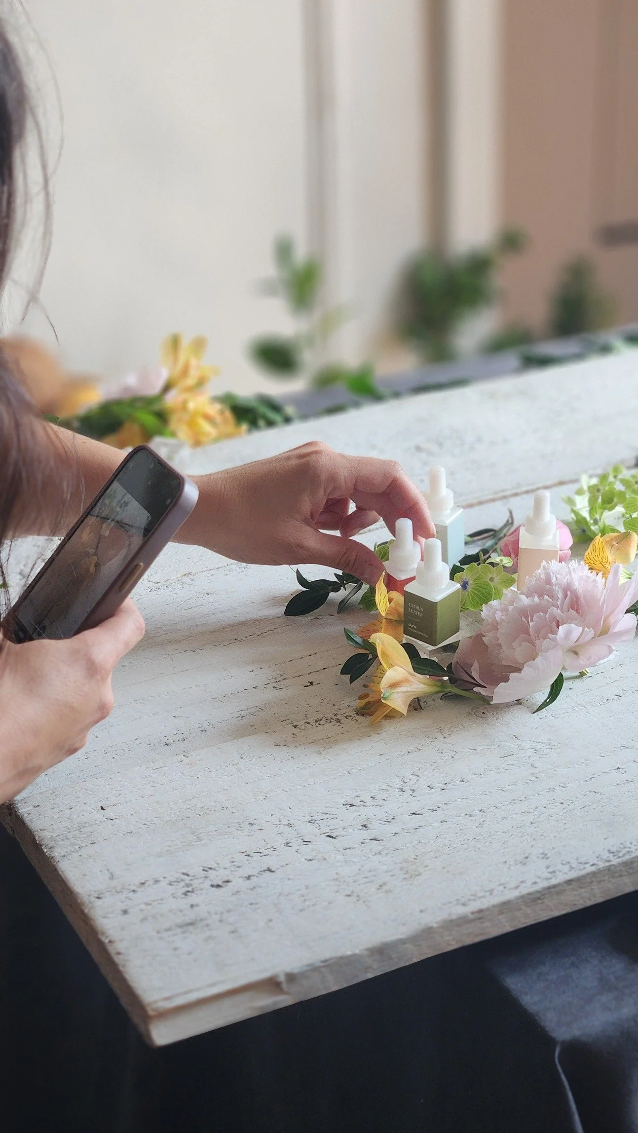 A person is taking a photo of small bottles of colorful liquids arranged with flowers on a rustic white table.