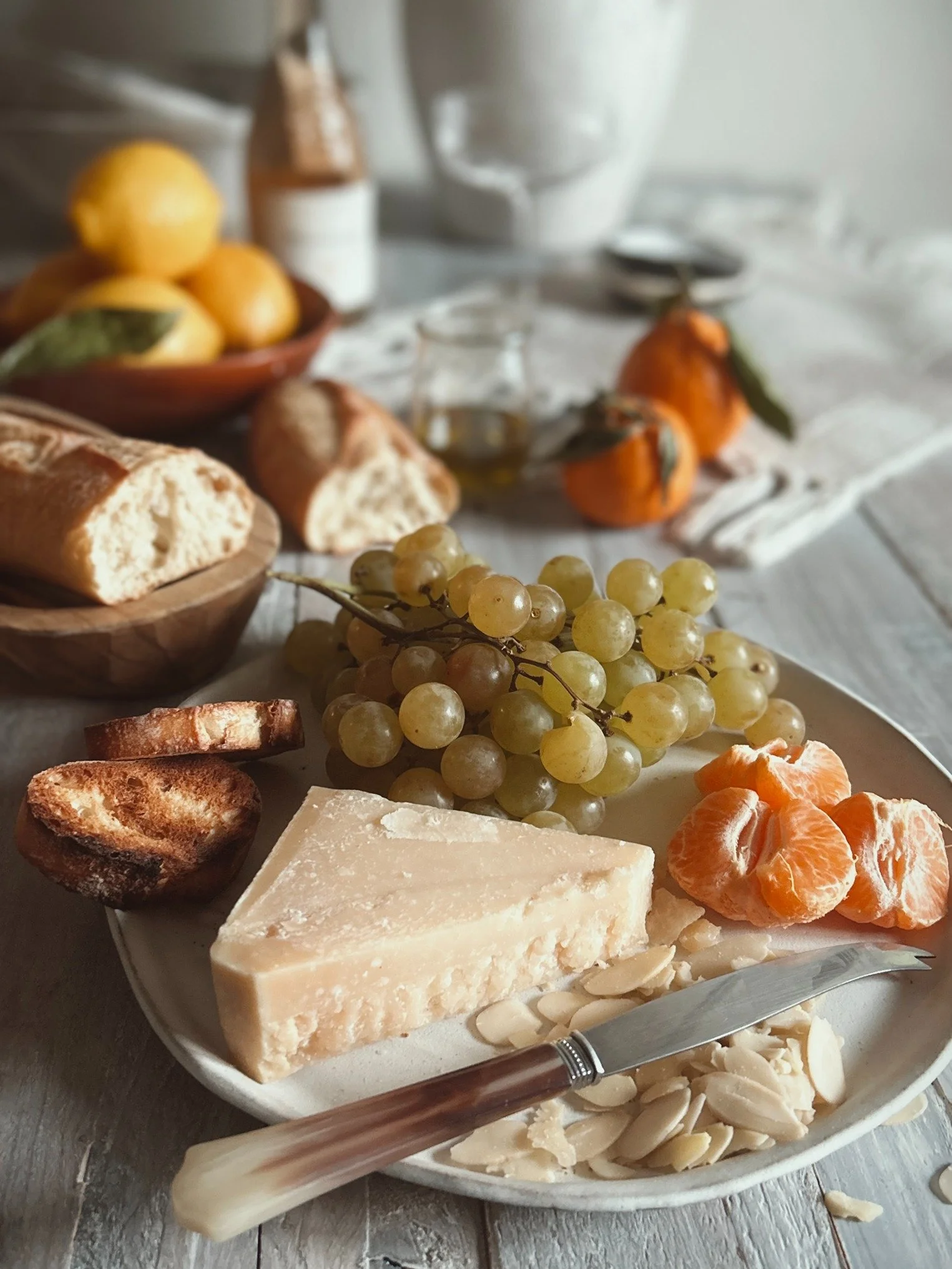 A cheese platter featuring a wedge of hard cheese, sliced almonds, tangerines, grapes, toasted bread, and a cheese knife on a white plate, with a rustic table setting including lemons, bread, and fresh fruits in the background.