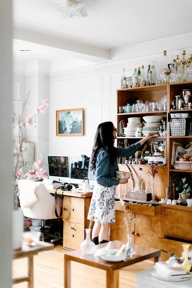 Woman looking at glassware on wooden shelf in a cozy, well-lit room with a computer desk, decorative items, and artwork.