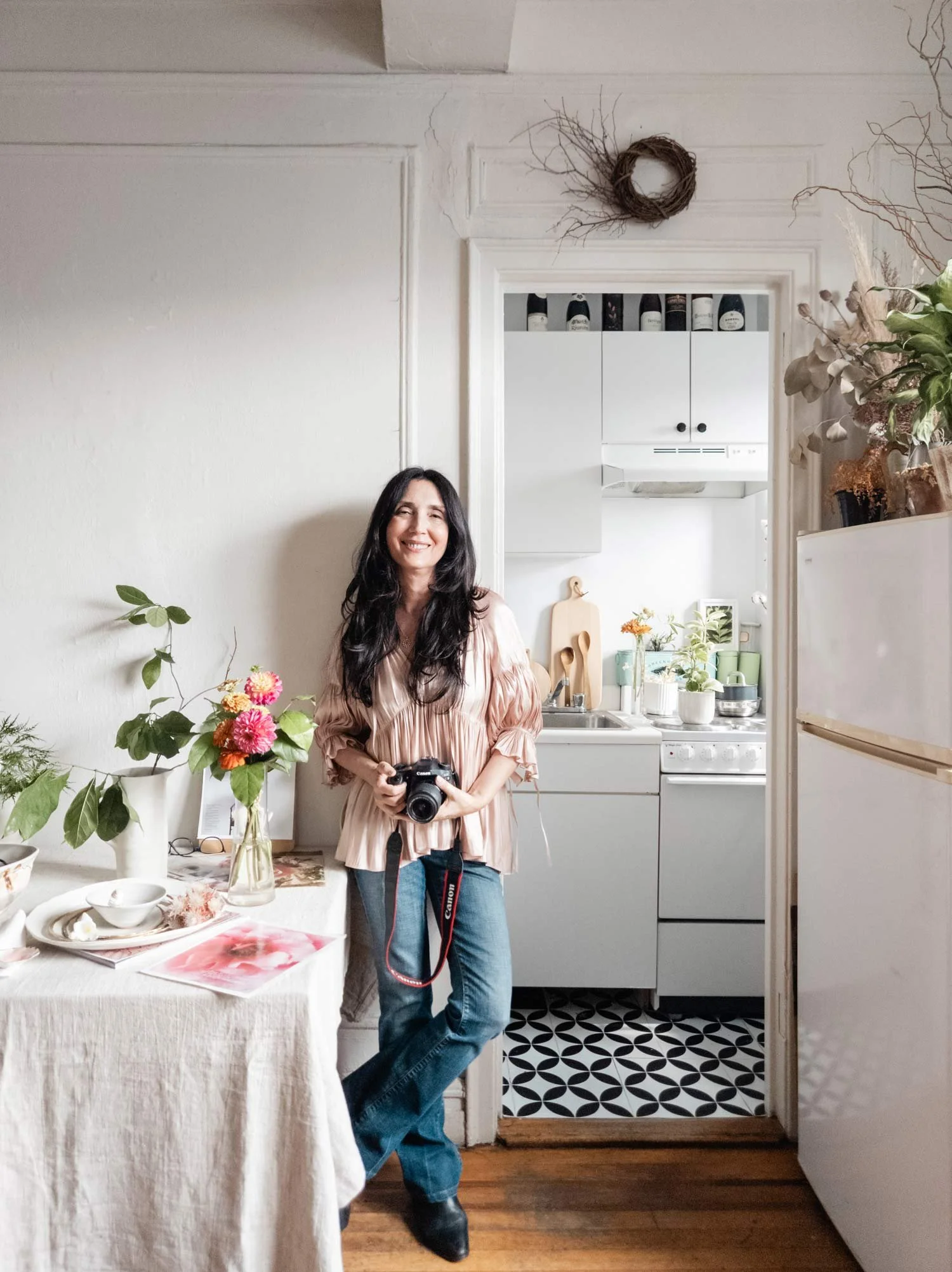 A woman with long dark hair standing in a cozy kitchen, holding a camera, smiling. The kitchen has white cabinets, a white fridge, and a small window. There are flowers on a table to her left and decorative branches above the door frame.