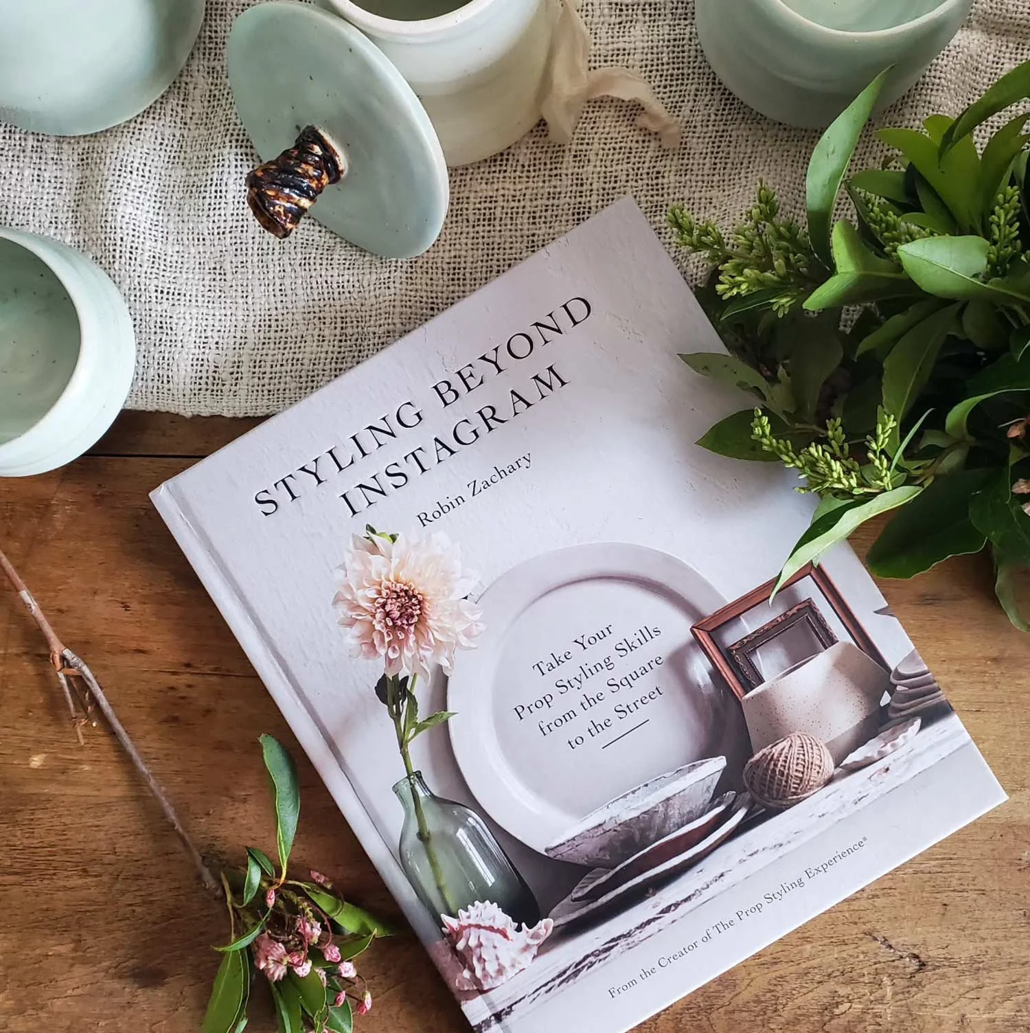 A book titled 'Styling Beyond Instagram' with a floral design on the cover, placed on a wooden table alongside a glass vase with pink flowers, green foliage, and ceramic dishes.