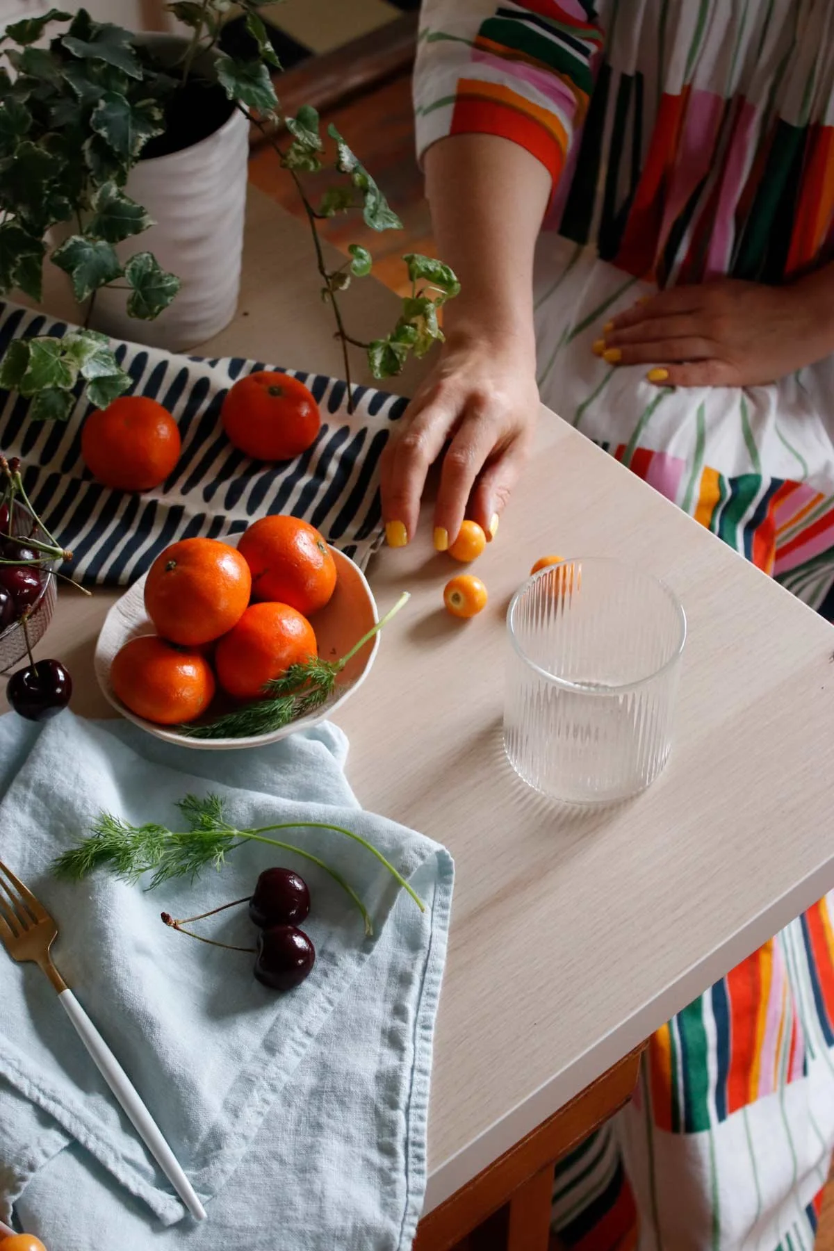A person dressed in a colorful striped outfit reaching for a small orange tomato on a table with various fruits including mandarins, cherries, and a glass of water.