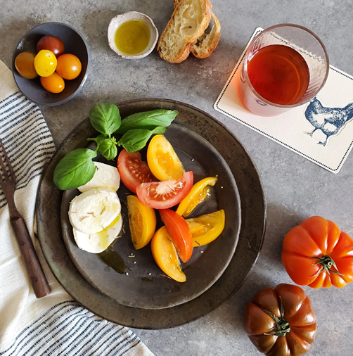 Caprese salad with sliced heirloom tomatoes, fresh basil, and mozzarella on a black plate. Surrounding the plate are a bowl of colorful cherry tomatoes, a small dish of olive oil, slices of toasted bread, a glass of tea, and fresh heirloom tomatoes on the table.