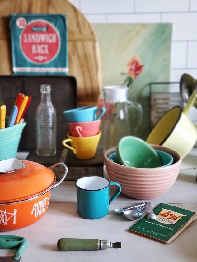 Kitchen countertop with colorful bowls, cups, utensils, and measuring tools, with a framed painting, a bouquet in a pitcher, and a bag of sandwich bags in the background.