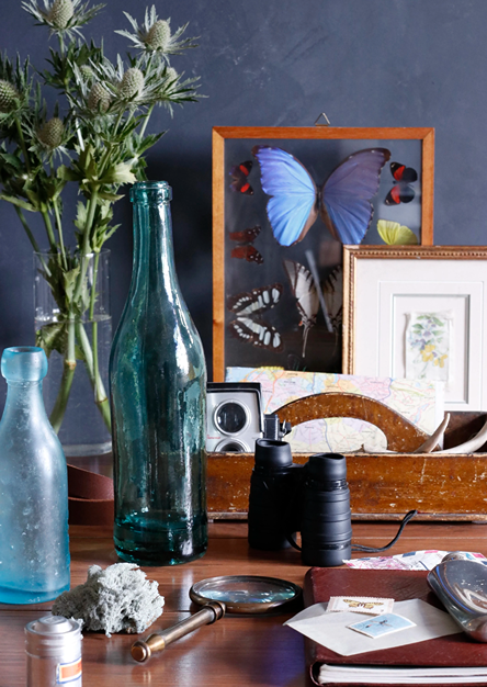 A cluttered table with vases, a large glass bottle, a pair of binoculars, framed butterfly artwork, and other decorative items.
