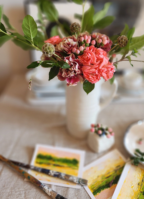 A white pitcher filled with pink and red flowers, including hydrangeas and greenery, on a table with watercolor paintings and a small decorated cake.