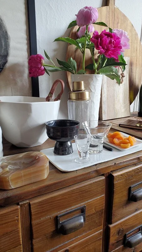 A wooden dresser holding a white bowl, a black chalice, a small shot glass, a clear glass bowl with an orange substance, a soapstone, and a white tray with a glass jar with a gold lid, a white ceramic vase with pink and purple peonies, several cutting boards, and other small objects.