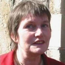 A woman with short brown hair wearing a red top, standing outdoors near a brick wall.