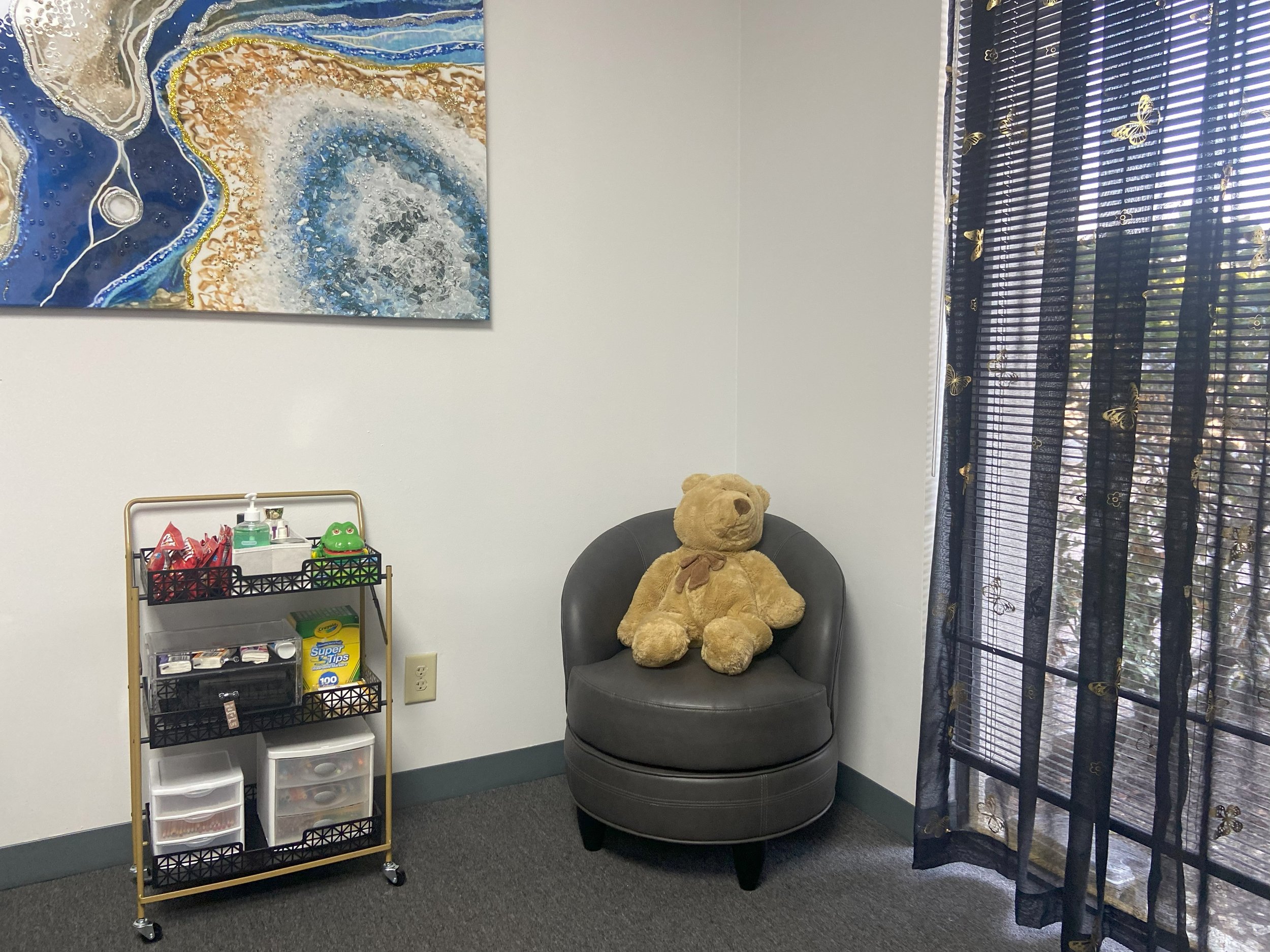 A small room with a black armchair and a large teddy bear sitting on it. To the left, there is a tiered storage cart filled with various office supplies, and below it, a plastic storage container. The wall features a colorful abstract painting, and a window with dark decorative curtains with butterfly and floral patterns allows natural light into the room.