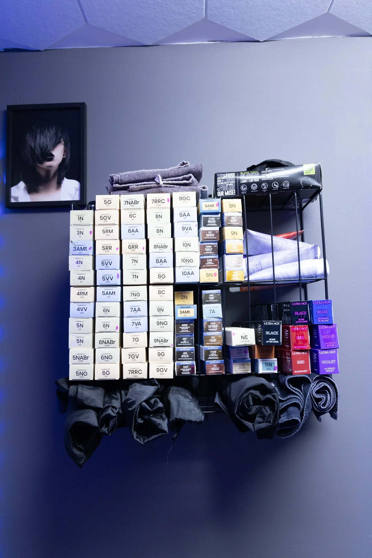 A wall-mounted display shelf with hair dye boxes, folded towels, and fabric rolls, with a framed black-and-white photo of a person with dark hair on a gray wall.