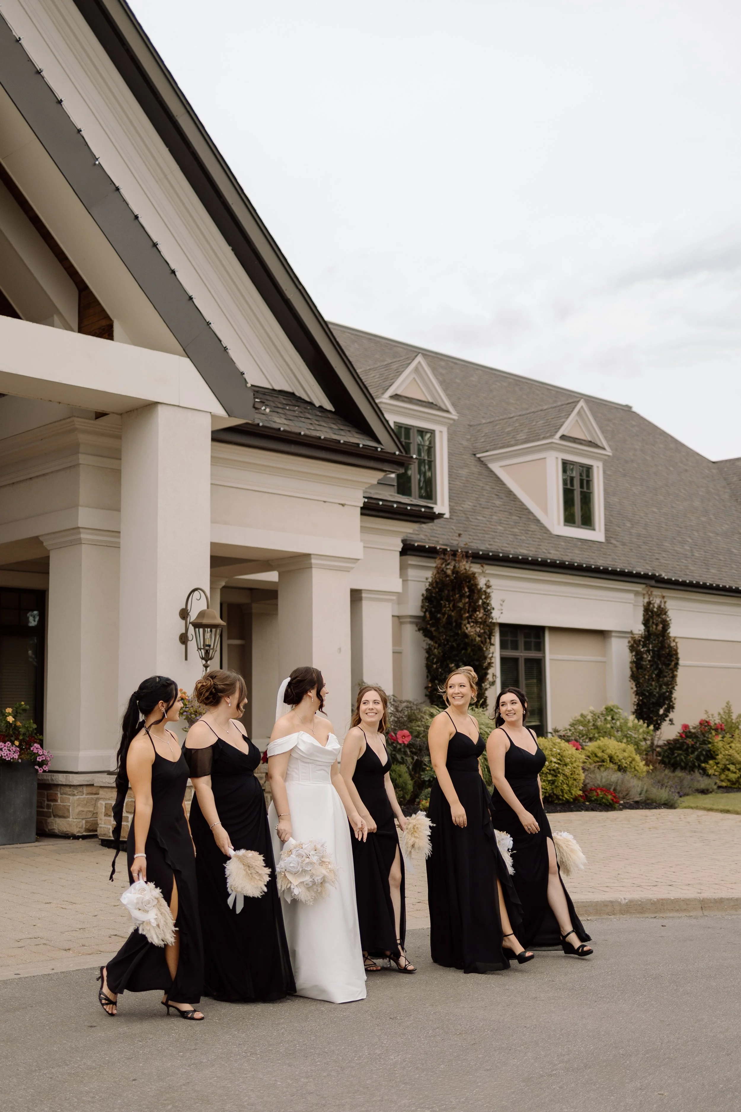 A bride and six bridesmaids walking outside in front of a large house with a gray roof and white siding. The bridesmaids are wearing black dresses, and the bride is in a white dress. They are holding bouquets and appear to be smiling.