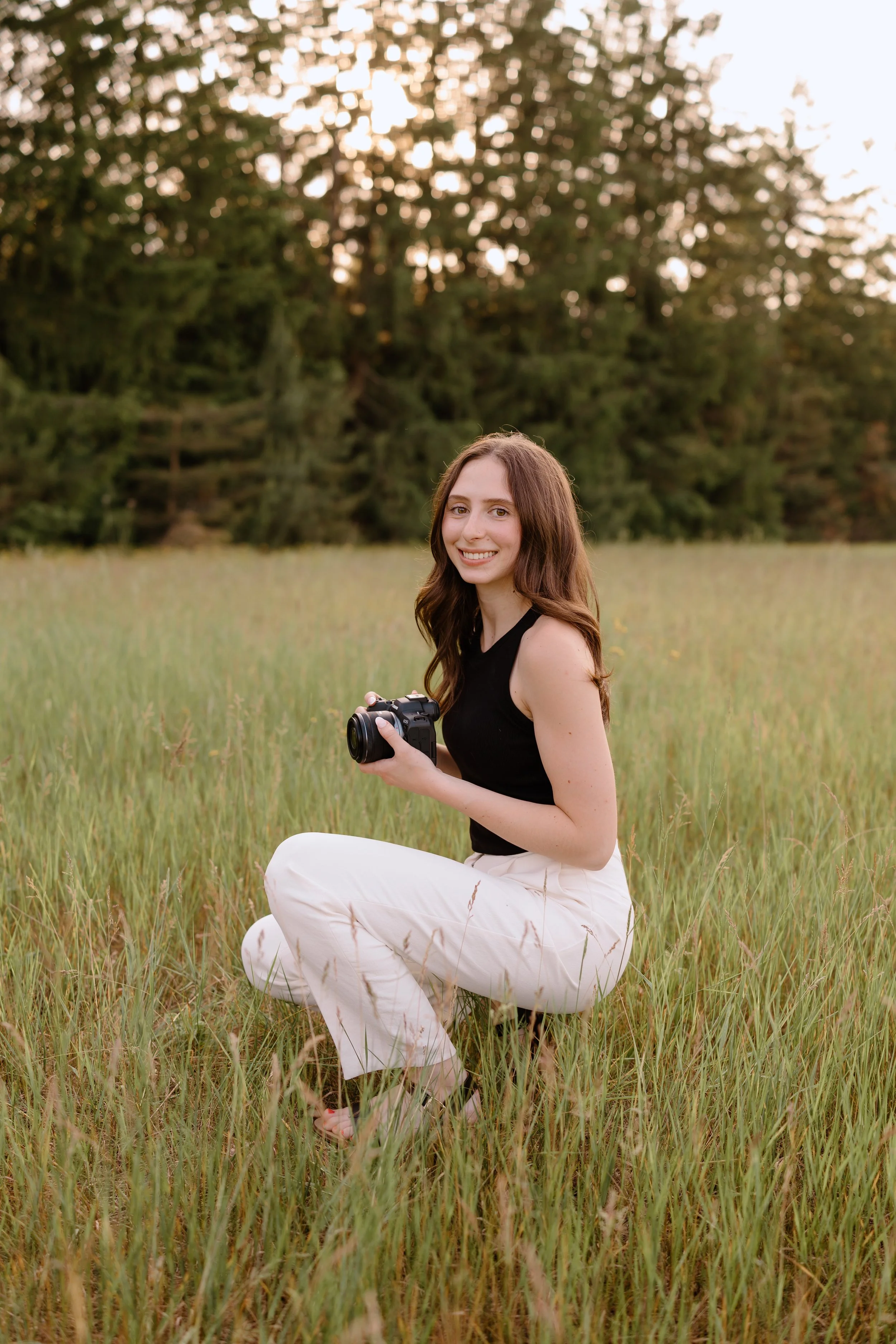 Photographer with brown hair crouching in a grassy field with professional camera in hand.